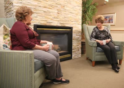 Two woman drinking coffee and sitting in chairs around a fireplace in memory care in pueblo